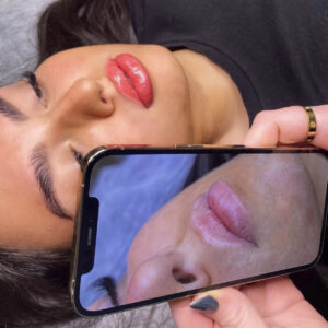 A model's lips are shown next to a phone with her before picture after having lip blush during a lip blush spmu course.